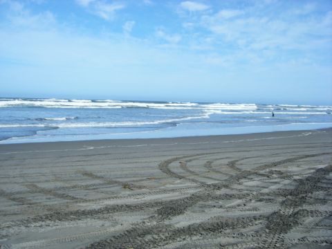 1994 Ford Explorer - Tire tracks are visible on the sand of a beach, with gentle waves rolling in from the ocean under a partly cloudy sky.