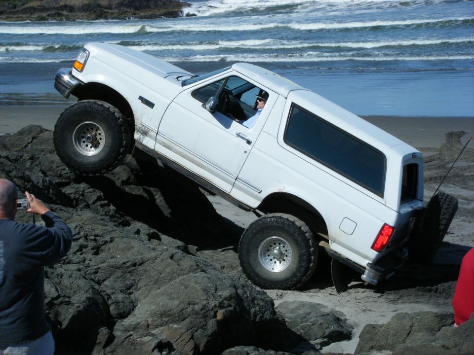 White SUV with large tires driving over rocks on a beach, ocean in background