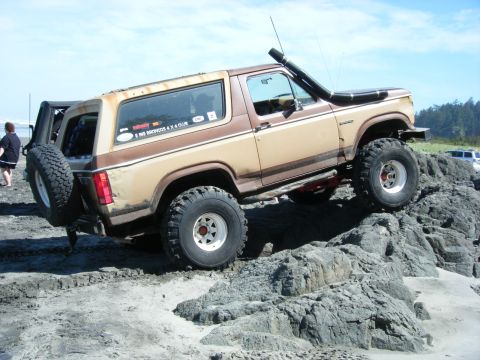 1994 Ford Explorer - A lifted Ford Bronco navigates rocky terrain on a beach, with another vehicle and people in the background.