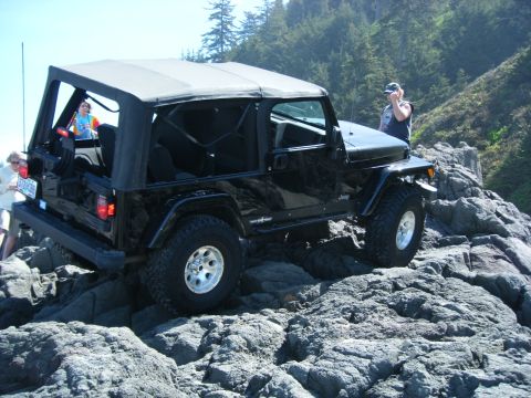 1994 Ford Explorer - A black Jeep Wrangler is shown off-roading on a rocky coastal area.