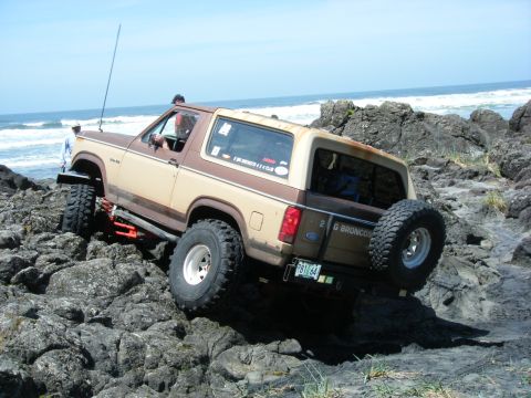 1994 Ford Explorer - This modified Ford Bronco is tackling a challenging rocky terrain near the coast, with the ocean waves in the background.