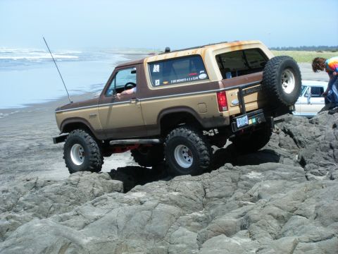 1994 Ford Explorer - A two-tone brown and tan Ford Bronco navigates rocky terrain by the ocean, with another vehicle visible in the background.