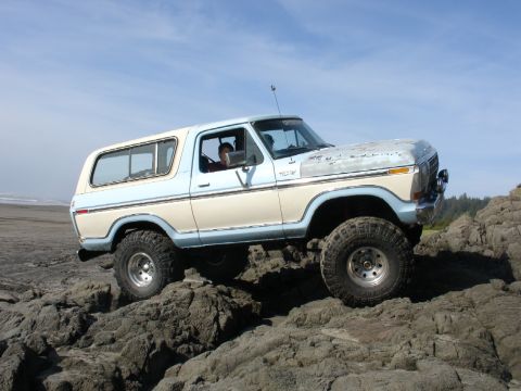 1978 Ford Bronco - A two-tone Ford Bronco navigates a rocky coastal terrain under a clear blue sky.