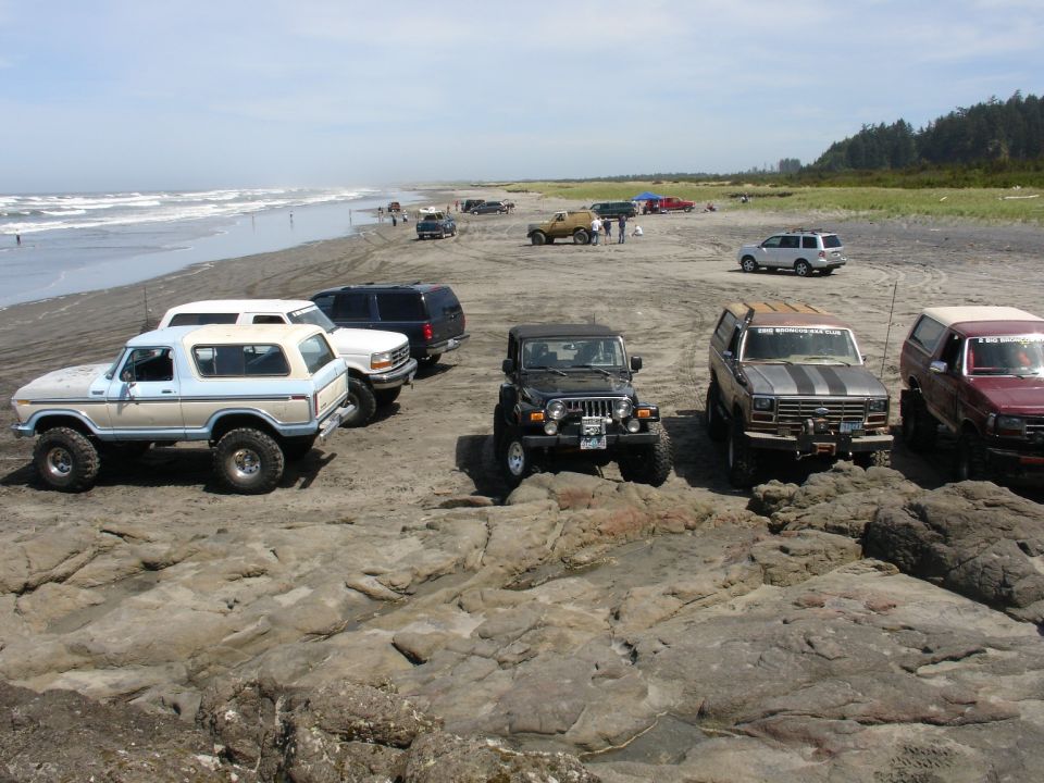 Several SUVs and Jeeps parked on a rocky beach near the ocean. One is a light blue and white Bronco