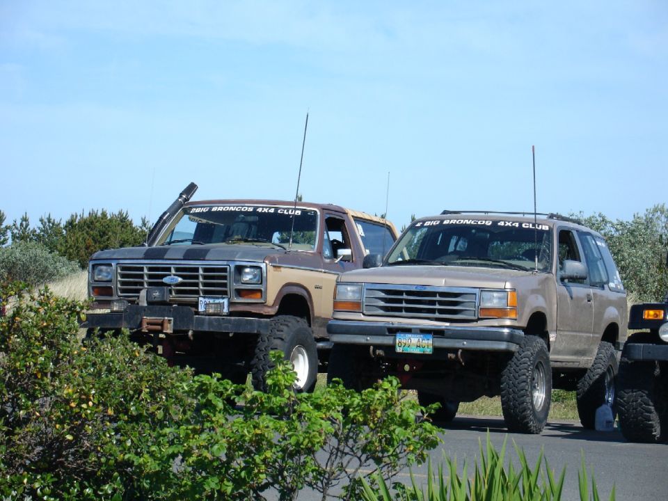 Two lifted SUVs, one a 1980-1986 Ford Bronco, the other a 1991-1994 Ford Explorer, parked off-road