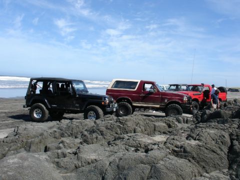 1978 Ford Bronco - A group of off-road vehicles, including a red 1978 Ford Bronco, are parked on a rocky beach under a blue sky.