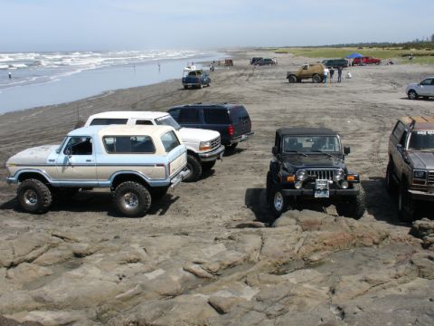 1978 Ford Bronco - Several off-road vehicles are parked on a beach near the ocean, with waves crashing in the background.