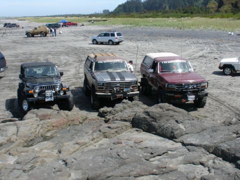 1978 Ford Bronco - Two SUVs, one maroon and one silver with racing stripes, are parked on a rocky beach with a black Jeep in the foreground.