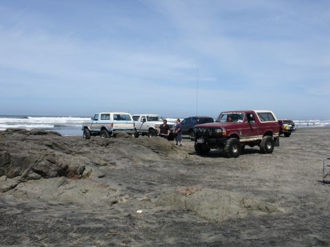 1978 Ford Bronco - Several SUVs, including a maroon 1978 Ford Bronco, are parked on a beach with waves in the background.