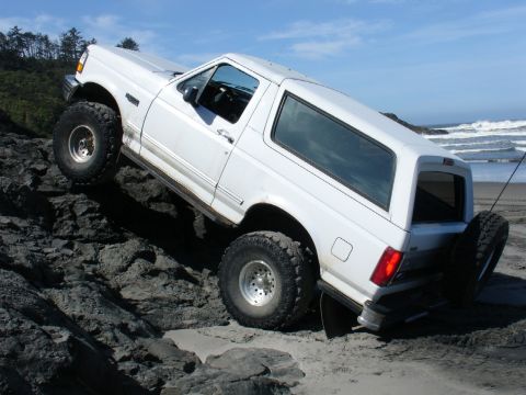 1978 Ford Bronco - A white Ford Bronco navigates a rocky incline on a sandy beach, with waves crashing in the distance.