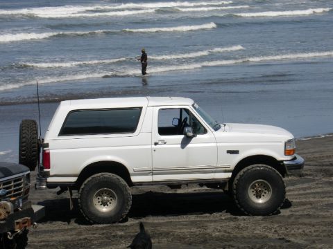 1978 Ford Bronco - A white Ford Bronco is parked on the beach near the ocean, with a person fishing in the surf.