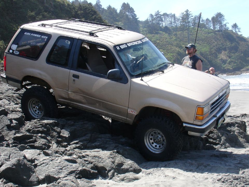 Tan Ford Explorer with lifted suspension and off-road tires on rocky terrain by the ocean. "2 BIG BRONCOS BEACH RUN" sticker on windshield