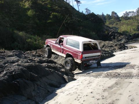 1978 Ford Bronco - A red and white Ford Bronco navigates a rocky shoreline on a sunny day.