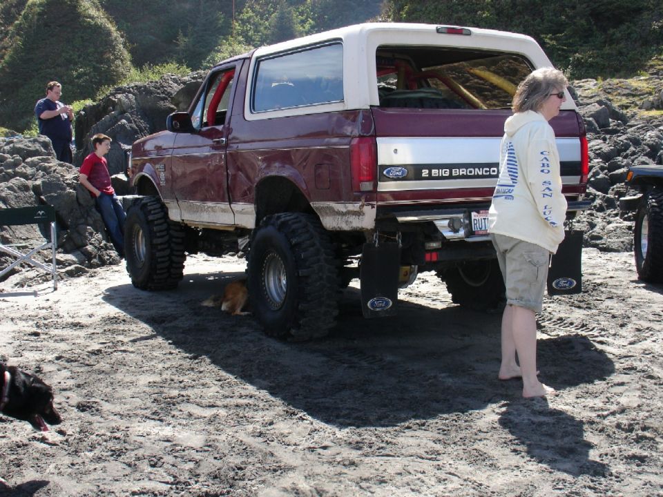 Maroon Bronco with lifted suspension and big tires on a sandy beach. Two people stand nearby