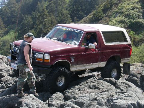 1978 Ford Bronco - A maroon Ford Bronco with a white top is shown off-roading on a rocky coastal path.