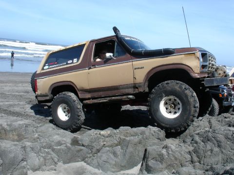 1978 Ford Bronco - A modified Ford Bronco navigates a rocky beach, with the ocean in the background and another vehicle nearby.