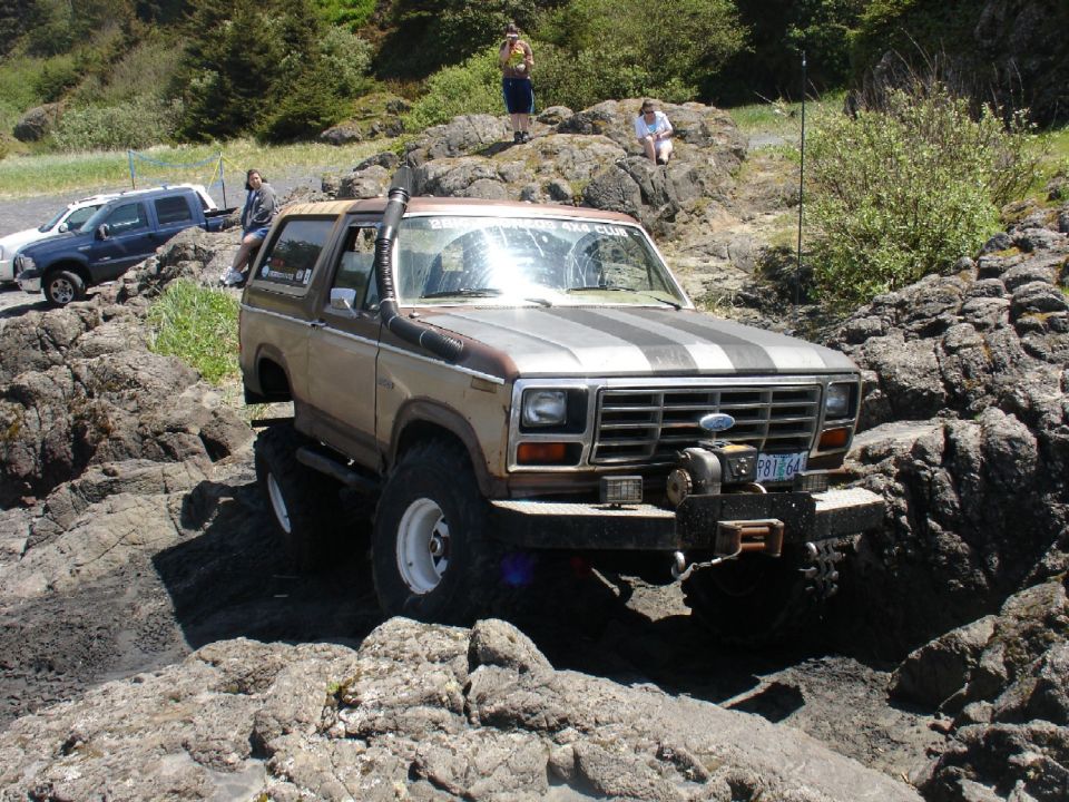 Tan and brown 1978 Ford Bronco with black hood stripes, lifted for off-roading over rocks