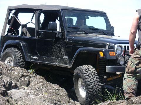 1978 Ford Bronco - A black Jeep Wrangler is shown off-roading over large rocks, with a person in camouflage pants standing nearby.