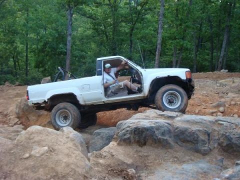 1978 Ford Bronco - A white pickup truck is shown off-roading over large rocks on a dirt trail.