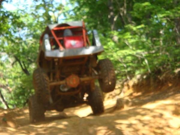 Off-road buggy with roll cage climbing a dirt hill, trees in background