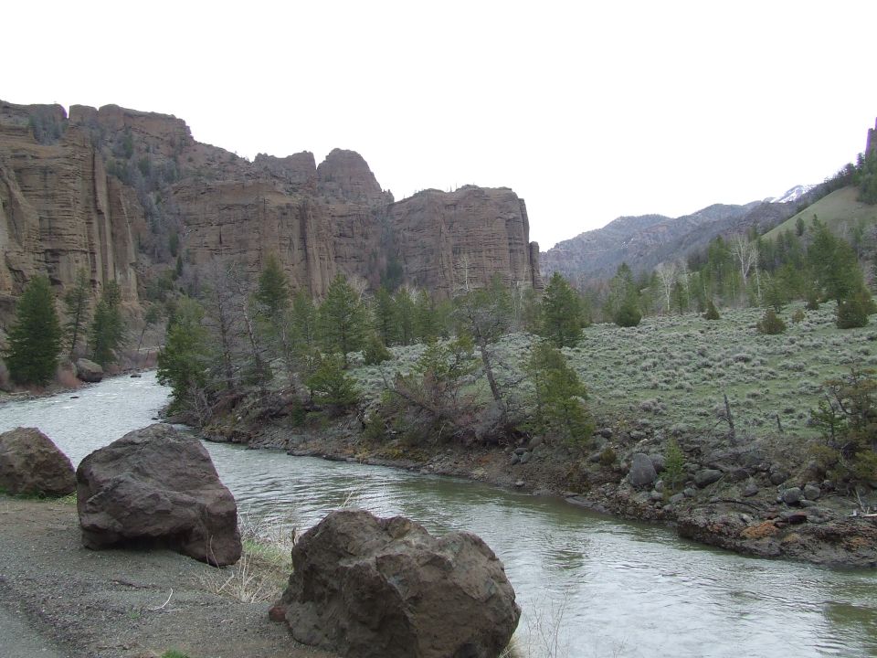 River flowing through a canyon with rocky cliffs and sparse trees under an overcast sky. Supermotors.net logo in lower right