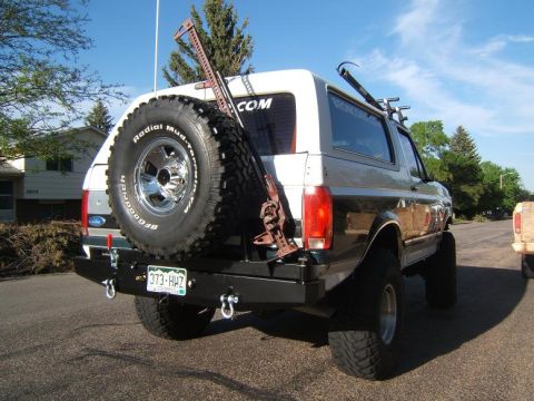 1993 Ford Bronco - This lifted Ford Bronco features a spare tire, a Hi-Lift jack, and a custom rear bumper, suggesting off-road modifications.