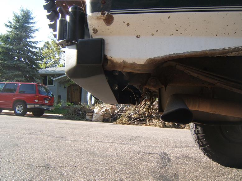 Close-up of a vehicle's rear bumper and exhaust pipe. A red SUV is parked in the background