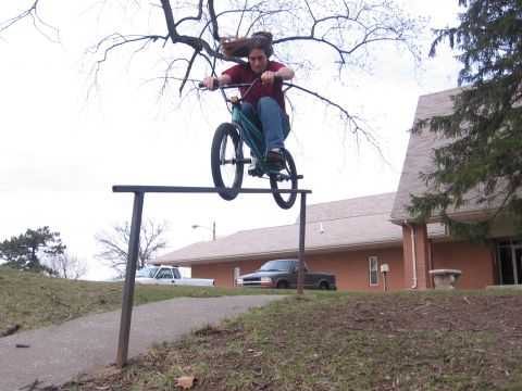 1988 Ford Bronco - A person is mid-air on a bicycle, performing a trick over a metal rail with buildings and trucks in the background.