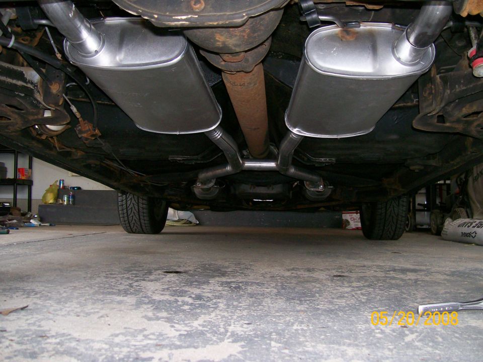 Underbody view of dual exhaust with two chrome mufflers and rusted driveshaft in a garage