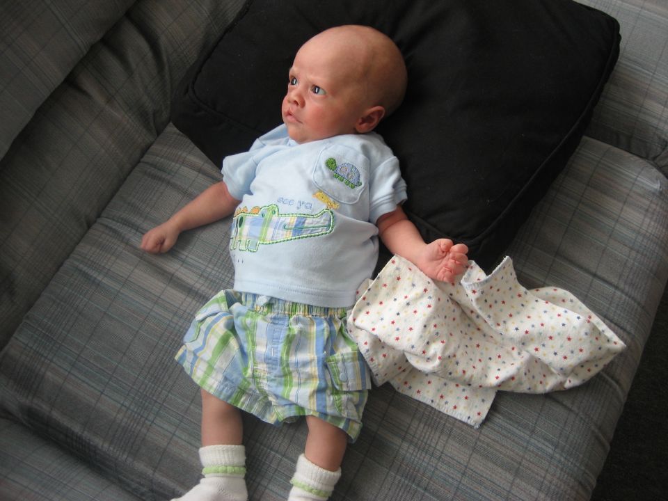 Baby lying on a couch, wearing a blue shirt with a cartoon alligator and plaid shorts