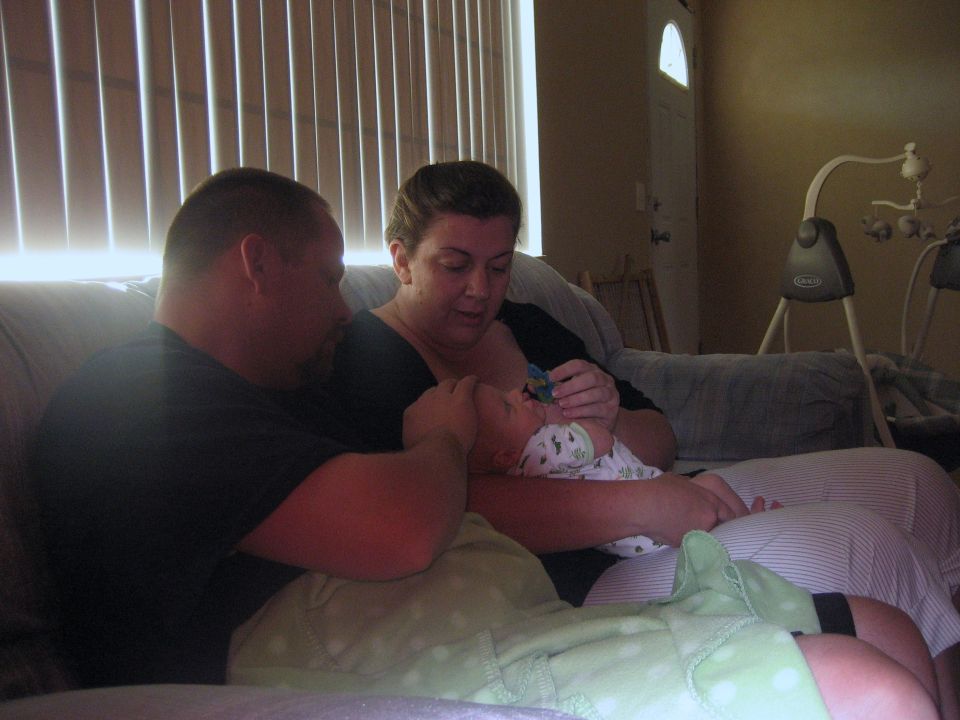 Man and woman on a couch holding a swaddled infant in a living room