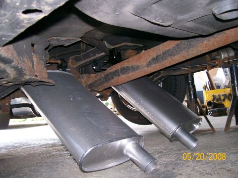 1995 Mercury Grand Marquis - Two new oval aluminized mufflers with offset inlet necks sit on the concrete floor beneath a car raised on jack stands, staged against the…