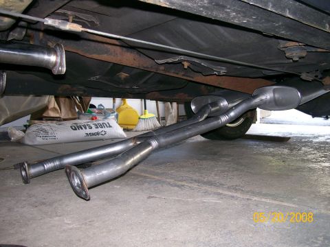 1995 Mercury Grand Marquis - A fabricated dual exhaust system laid out on the garage floor beside a car raised on stands, showing two parallel pipes with flared flange…