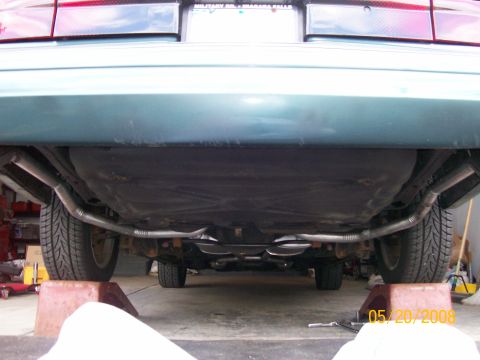 1995 Mercury Grand Marquis - Low rear view under a dark green sedan up on ramps in a garage, showing a newly fabricated dual exhaust with two chrome tips exiting side b…
