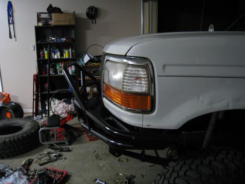1996 Ford Bronco - Front driver-side corner of a white 1990s Ford truck sitting in a garage mid-project, with a black tube bumper and brush guard fitted below…