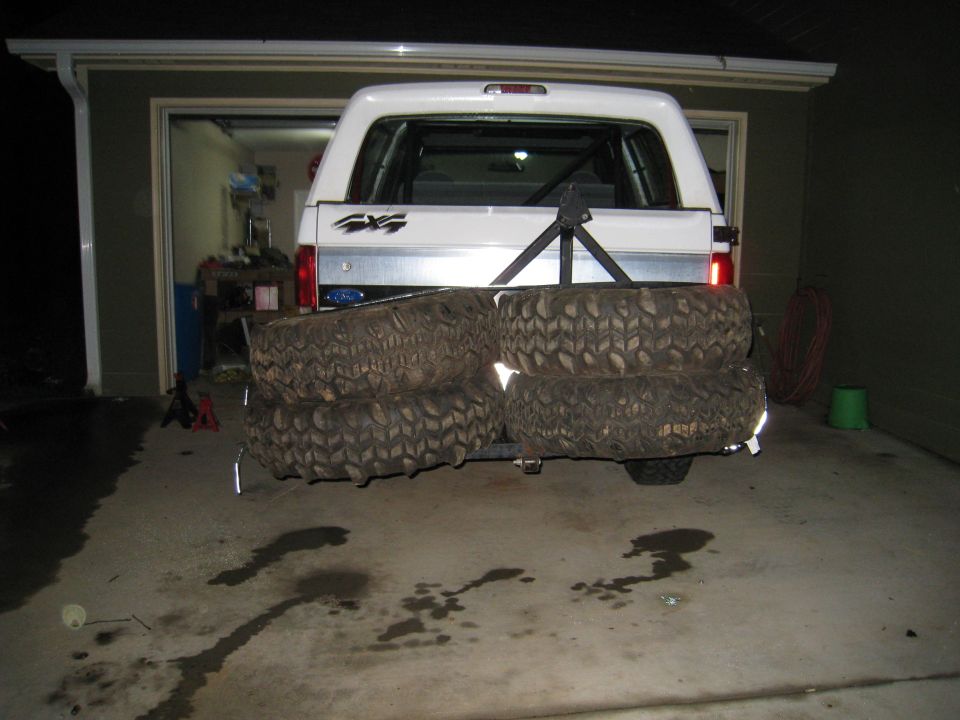 Rear view of white Ford 4x4 SUV in garage with four muddy mud-terrain tires stacked behind it