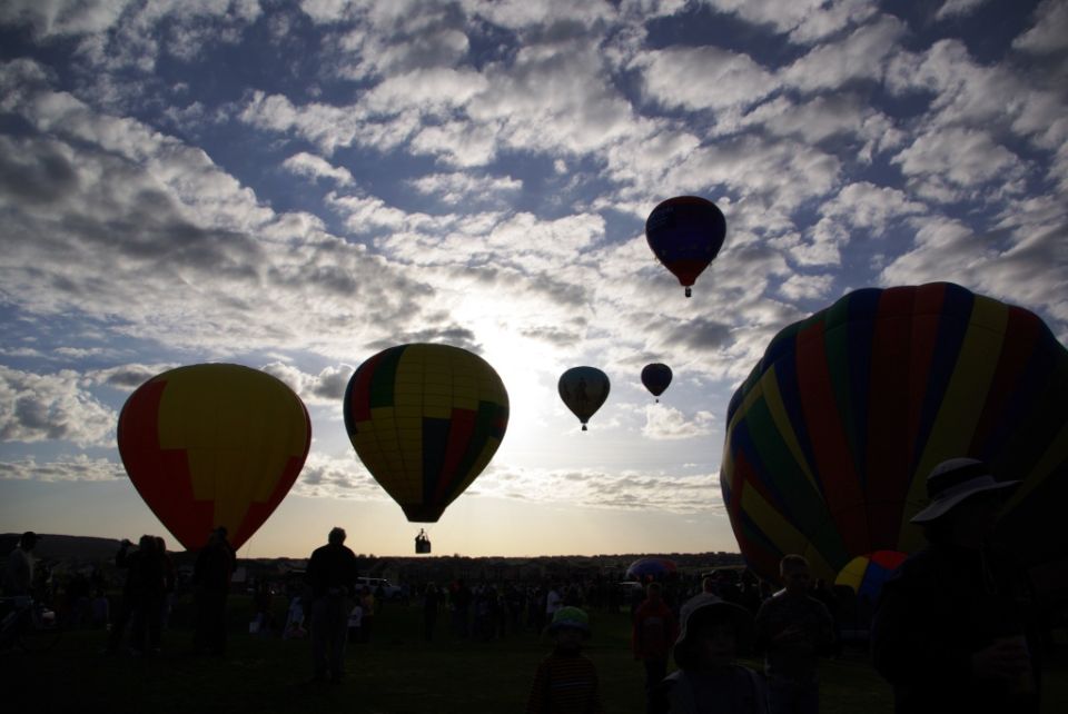 Several hot air balloons ascend into a cloudy sky over a crowd