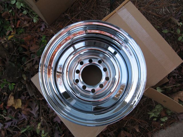 Chrome steel wheel with eight lug holes sitting on a cardboard box on leaf-covered ground