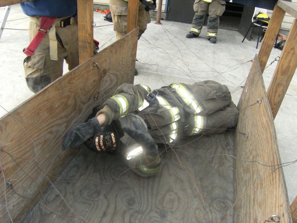 Firefighter in turnout gear lying on plywood floor within a wooden structure, tangled in wires