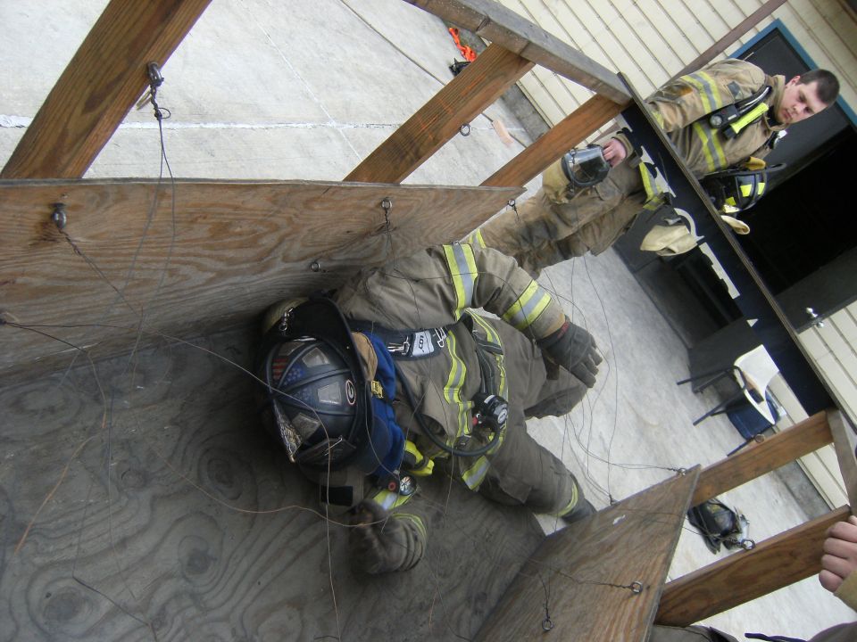 Firefighters in protective gear practice maneuvering through a wooden structure, with one firefighter on the ground and another observing from above