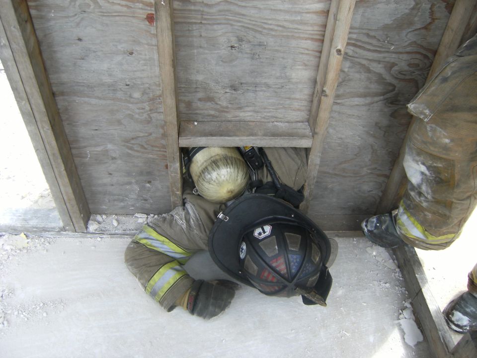 Firefighter in protective gear crawling through a wooden opening, wearing a helmet and carrying an oxygen tank