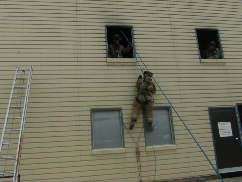 Firefighters rappelling down a building. One is descending a rope from a window, while others are visible in upper windows
