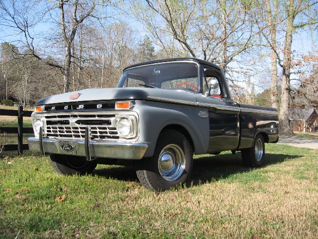 Black and gray-primer 1960s Ford F-100 pickup on a lawn by a split-rail fence