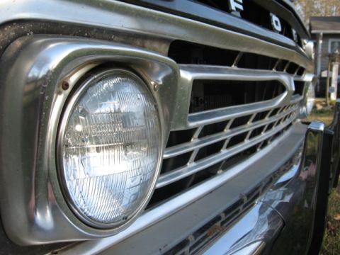 1966 Ford F-100 - Tight close-up of the driver-side round sealed-beam headlight and chrome bezel on a vintage Ford pickup, with the horizontal-bar aluminum g…