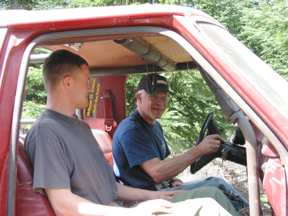Two men in the cab of a red vehicle on a wooded trail, driver at the wheel with a CB mic hanging overhead