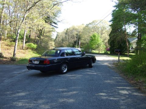 1999 Ford Crown Victoria - A dark sedan is parked on a residential street with trees and a house visible.