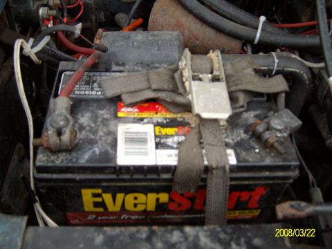 1988 Ford Bronco - Overhead view of a black EverStart battery sitting in a dirty engine bay, held down by a makeshift gray nylon strap and ratchet buckle inst…