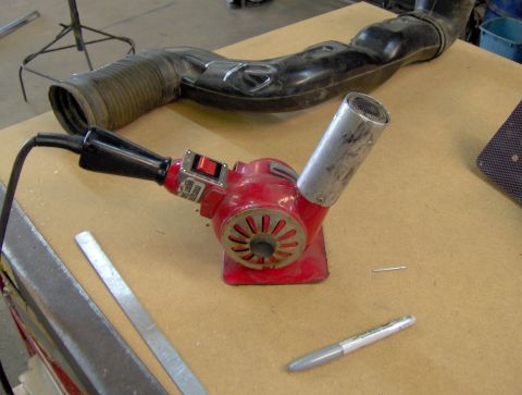 1978 Ford Bronco - A red heat gun and a vehicle air intake hose are laid out on a workbench, along with a marker and a small metal object.