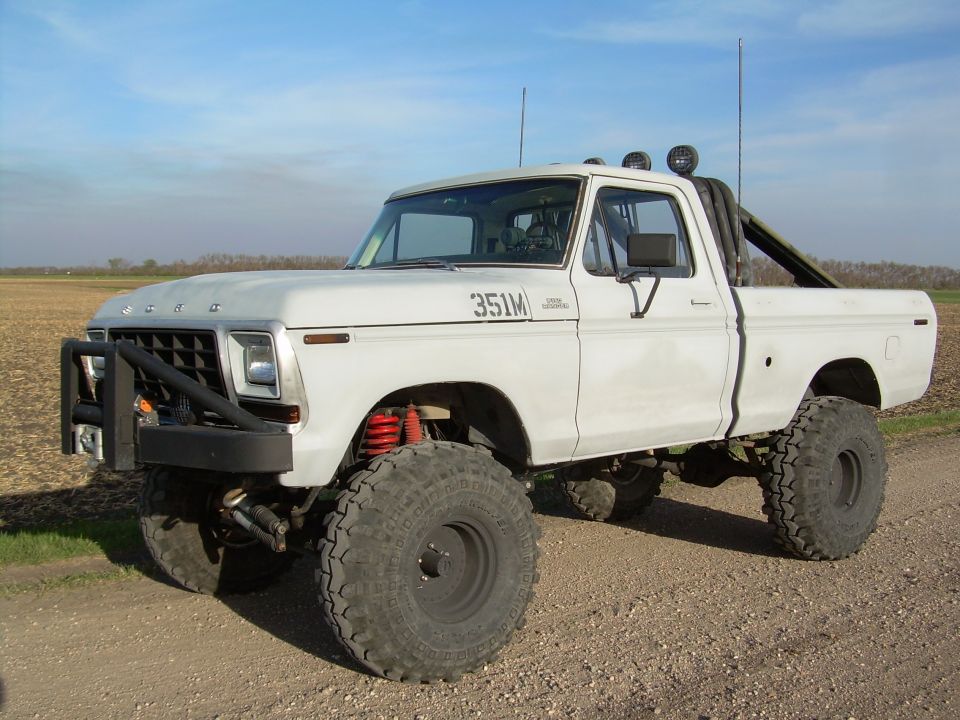 Lifted white Ford F-150 pickup with 351M hood lettering, big off-road tires and light bar, on a gravel road.