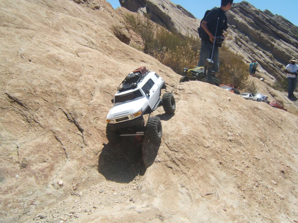 White RC rock crawler climbing a rocky hill, person with controller in background